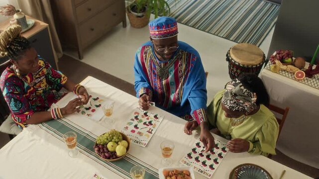 High angle view of African American young man helping Black female friend put last card on printed sheet and they clapping hands to celebrate her win when playing tabletop game on Kwanza day
