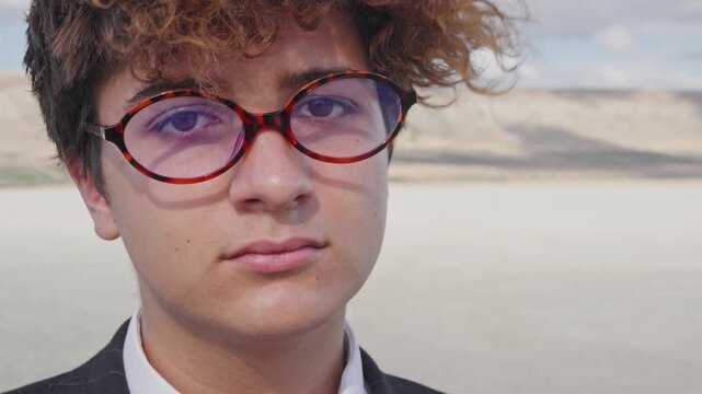 Portrait of a curly-haired teenager with glasses and a mature business suit against a backdrop of a hill and blue sky. The camera pans.
