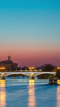 Seville's skyline day to night transition timelapse featuring the Torre Sevilla skyscraper near Guadalquivir River. The illuminated Triana Bridge reflects on water, modern and traditional architecture
