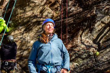 Naklejka premium Rock climber (young woman, in blue helmet, hardshell jacket, and blue climbing harness) standing in front of a rock face with belay line hanging vertically.