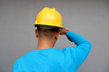 Back view of a person wearing a bright yellow hard hat and blue t-shirt standing against a neutral gray background
