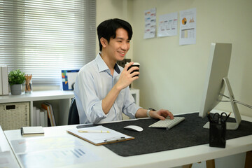 Professional Asian man enjoying a coffee break while looking at a computer in a relaxed office atmosphere
