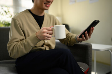 Casual morning vibe with cropped shot of a young man holding a white mug and looking at his phone, enjoying peaceful downtime