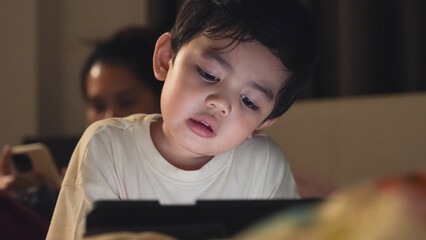 A young boy intensely focused on a digital tablet in a dimly lit bedroom, highlighting screen time and modern parenting.