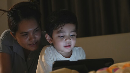 A young boy intensely focused on a digital tablet in a dimly lit bedroom, highlighting screen time and modern parenting.
