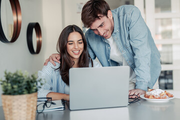 A young couple enjoys a morning at home, smiling while working together on a laptop. The warm light and teamwork reflect love, productivity, and a modern lifestyle balance.