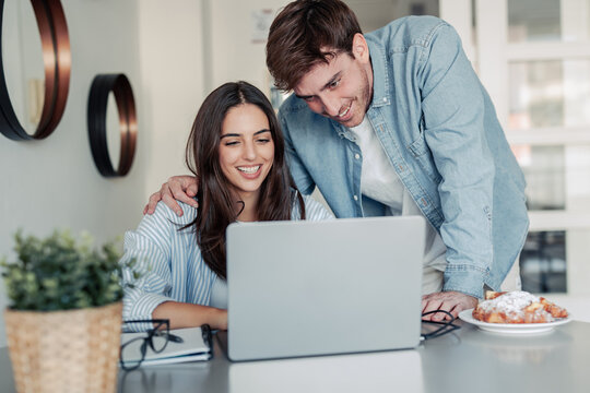 A young couple enjoys a morning at home, smiling while working together on a laptop. The warm light and teamwork reflect love, productivity, and a modern lifestyle balance.
