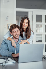 A smiling woman stands behind her partner as he works on a laptop at home. Their teamwork and shared focus reflect motivation, love, and the balance of modern relationship goals.