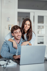 A smiling woman stands behind her partner as he works on a laptop at home. Their teamwork and shared focus reflect motivation, love, and the balance of modern relationship goals.
