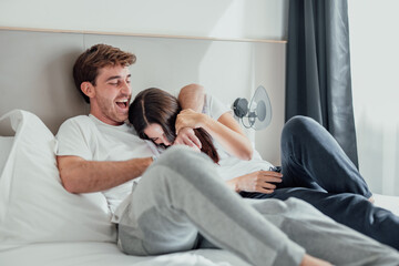 A young couple lies in bed, laughing and playfully teasing each other. Their smiles and playful...