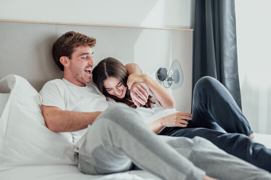 A young couple lies in bed, laughing and playfully teasing each other. Their smiles and playful touches show joy, closeness, and love, sharing a fun, relaxed moment at home.