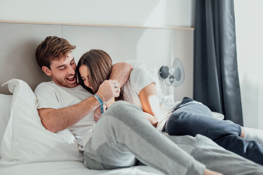A young couple lies in bed, laughing and playfully teasing each other. Their smiles and playful touches show joy, closeness, and love, sharing a fun, relaxed moment at home.