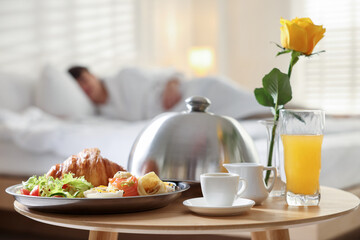 Delicious breakfast served on wooden table and man lying in bed at hotel room, selective focus