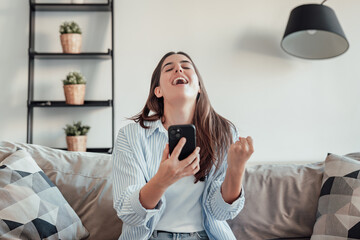 A young woman sits on the sofa, holding her smartphone with excitement and joy. She celebrates a special offer, lottery win, or dream opportunity, feeling victorious and overjoyed.