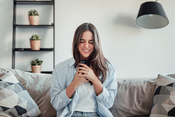 A young woman sits on the sofa, holding her smartphone with excitement and joy. She celebrates a special offer, lottery win, or dream opportunity, feeling victorious and overjoyed.
