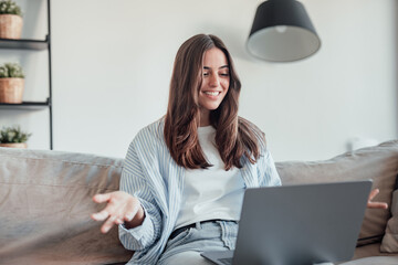 Smiling girl sit on couch at home wave talk with friend or family having video call on laptop,...