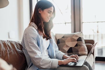 Concentrated millennial girl sit on couch working on laptop browsing internet at home during lazy weekend, focused young woman freelancer busy using computer surfing wireless web shopping online