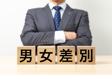 「男女差別」と書かれたブロックと腕組みをするビジネスマン