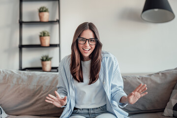 Obraz premium A young woman sits on the sofa wearing glasses, smiling during a video call. She enjoys chatting online, recording her vlog, and sharing happy, cheerful moments from her modern home.