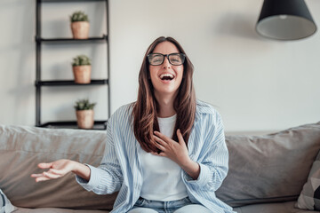Obraz premium A young woman sits on the sofa wearing glasses, smiling during a video call. She enjoys chatting online, recording her vlog, and sharing happy, cheerful moments from her modern home.