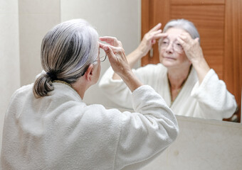 Senior woman looking at her face in the bathroom applying anti age cream. Elderly person wearing a...