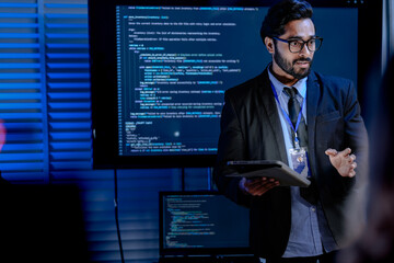 An IT support male engineer dressed smartly with a headset actively assists a user analyzing system data on his computer in a dimly lit control room setting ensuring smooth operations.