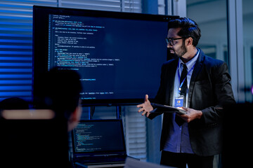 A visionary male AI programmer in a futuristic setting with a headset actively presents complex code and data on a large screen discussing advanced machine learning algorithms.