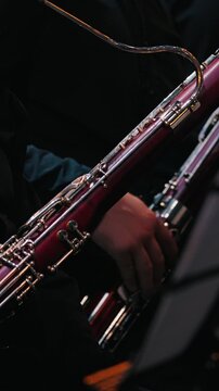 A close-up of a musician's hand holding a bassoon, revealing its intricate keys and polished wood. The brass section of an orchestra