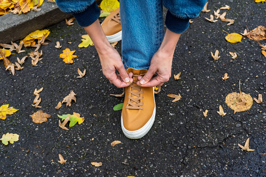 A person in stylish blue jeans tying the laces of their trendy tan sneakers on an asphalt path, surrounded by vibrant fallen autumn leaves, symbolizing readiness for a seasonal walk