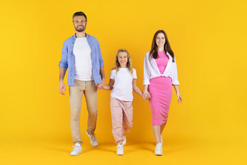Fototapeta premium Smiling family walking together in studio on yellow background. Parents and daughter holding hands, dressed in bright colors, expressing joy and unity, copy space