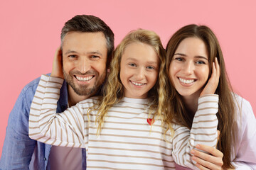 Happy family of three smiling in studio, daughter touching parents faces playfully. Pink background adds warmth and tenderness to joyful family portrait
