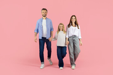 Fototapeta premium Young family of three walking hand in hand in studio, pink background. Parents and daughter smiling confidently, symbolizing unity and happy lifestyle together
