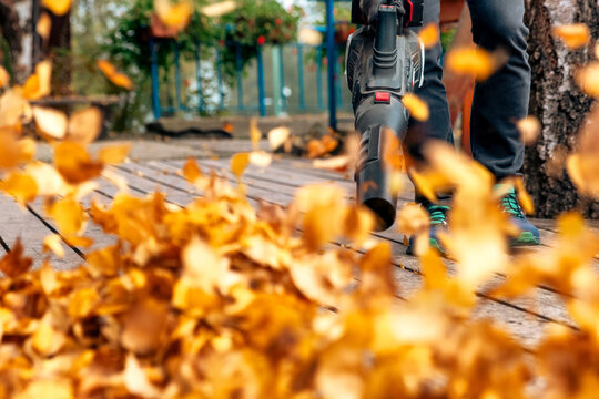 Man using handheld blower to remove dry leaves from wooden terrace during autumn yard cleaning