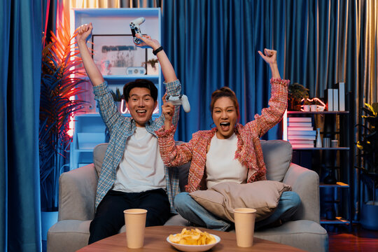 Couple gamer raising fist up holding joysticks be winner together complete level sitting sofa playing video game with front snack and drinks at neon light color living room at modern home. Infobahn.