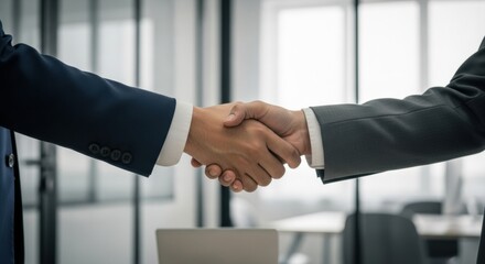 Two businessmen in navy and grey suits shaking hands to confirm a partnership in a contemporary office with glass partitions in the background.