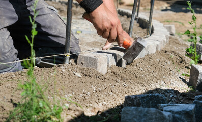 Landschaftsg&auml;rtner bei der Arbeit im Detail &ndash; pr&auml;zise Handarbeit im Gartenbau mit Fokus auf Steine, Werkzeug und Struktur. Selektive Sch&auml;rfe, viel Copy Space