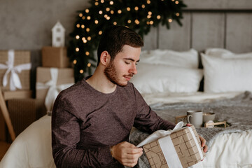 Young man in a casual sweater sitting in a cozy bedroom, carefully unwrapping a Christmas gift wrapped in brown paper and white ribbon, with blurred holiday decor in the background