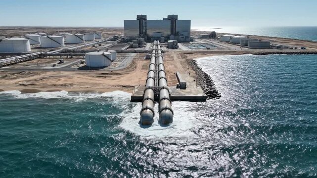 Industrial water desalination plant operates with large pipelines extending toward the ocean. Bright sunny day at coastal location highlights advanced technology