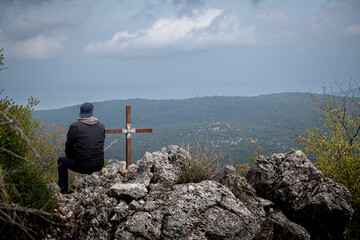 Photo of a man wearing cap and hoodies sitting  on a rock next to an iron cross watching the view in the mountains.