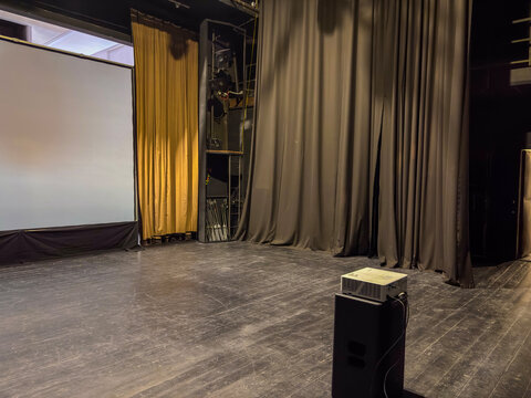 interior of a small, dimly lit theater stage or backstage area with a black wooden floor, heavy curtains, a white projection screen, and a projector on a stand
