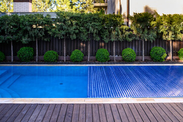 A partially covered outdoor swimming pool with a blue slatted pool cover in a private backyard. The area is surrounded by well-maintained shrubs, conifer trees, and a dark wooden fence