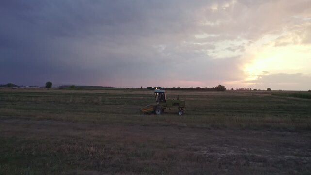 A farmer on a tractor with a machine that collects the hay in rows.
Tractor Working on Farm at Sunset Time. 