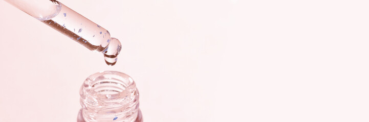 Close-up of dropper with translucent liquid above open bottle against soft pink background.