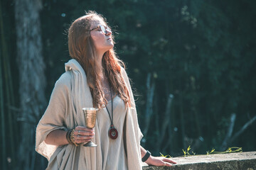 Portrait of a serene woman in a linen hooded dress holding an ornate metal goblet in a lush garden, enjoying the sunlight.