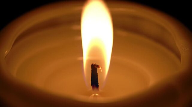 A lit candle with a tall bright flame burning above the dark wick in a pool of wax