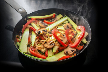 Stir fried vegetables in a steaming frying pan with red bell pepper, zucchini, mushrooms and onion on a black induction hob, vegetarian cooking concept, copy space, selected focus