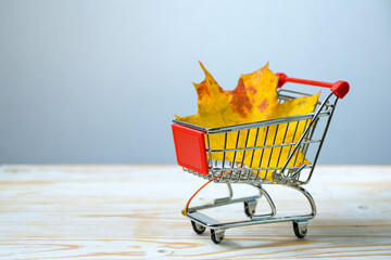 Autumn sale, shopping cart with an orange-yellow maple leaf on light wood against a gray-blue background, seasonal advertisement concept, copy space, selected focus