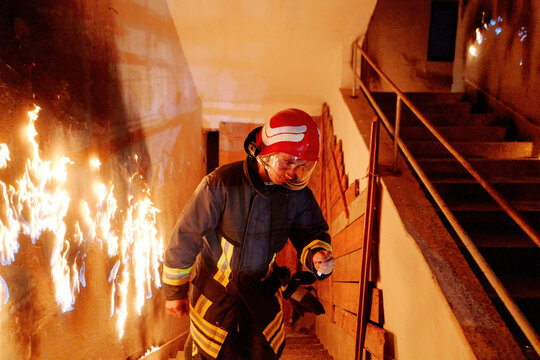 Firefighter in action, battling flames in burning building, wearing protective gear and helmet, fire safety and emergency response, danger and rescue, urban environment.