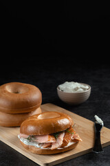 A bagel with salmon and cream cheese is on a wooden cutting board. A bowl of cream cheese is next to the sandwich