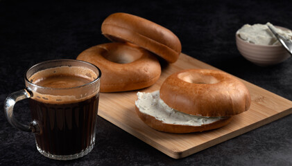 A glass of coffee and two bagels on a wooden board. The coffee is black and the bagels are brown